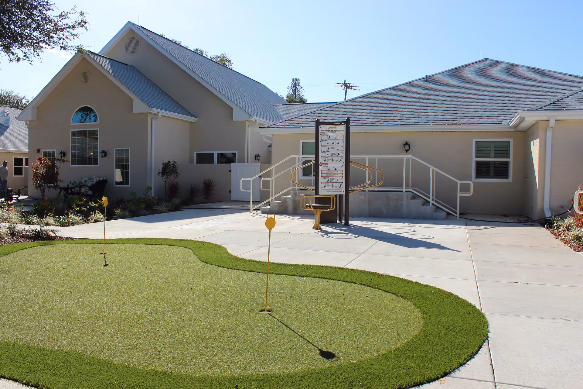 Exterior view of a beige senior care building with a small artificial putting green, concrete walkway, and an accessible ramp with an exercise sign.