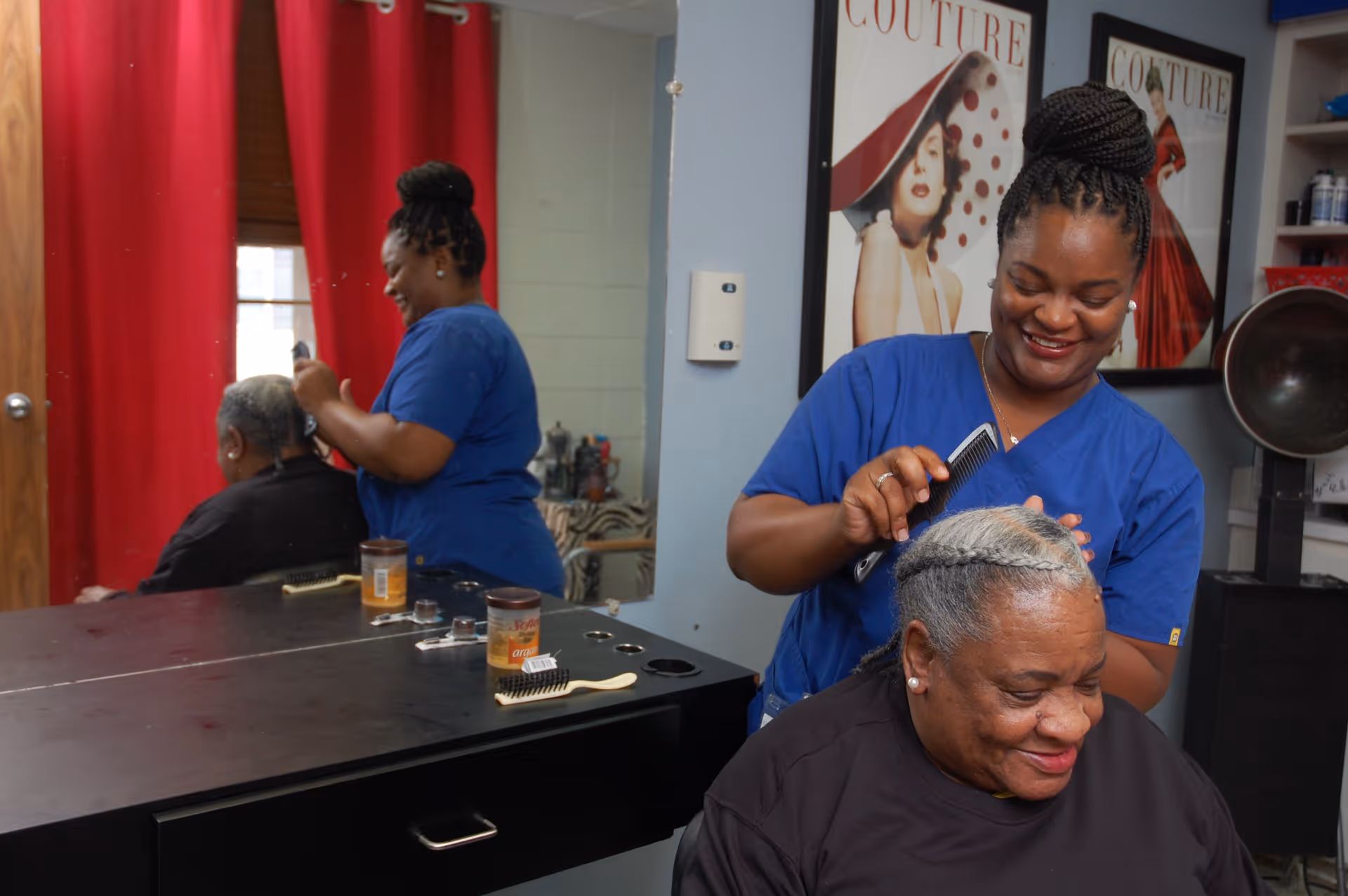 A hairstylist in blue scrubs is smiling while combing the hair of an elderly woman seated in a salon chair. The salon has a large mirror, red curtains, and framed fashion posters on the wall.