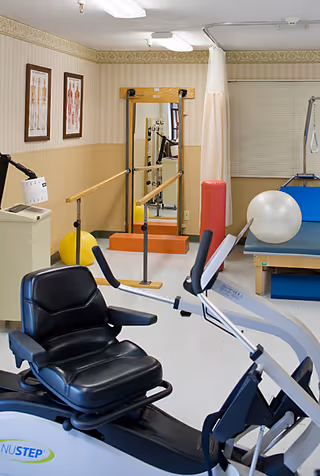 Rehabilitation/exercise room with a recumbent exercise bike in the foreground, parallel bars, therapy table, exercise balls, and a wall mirror.