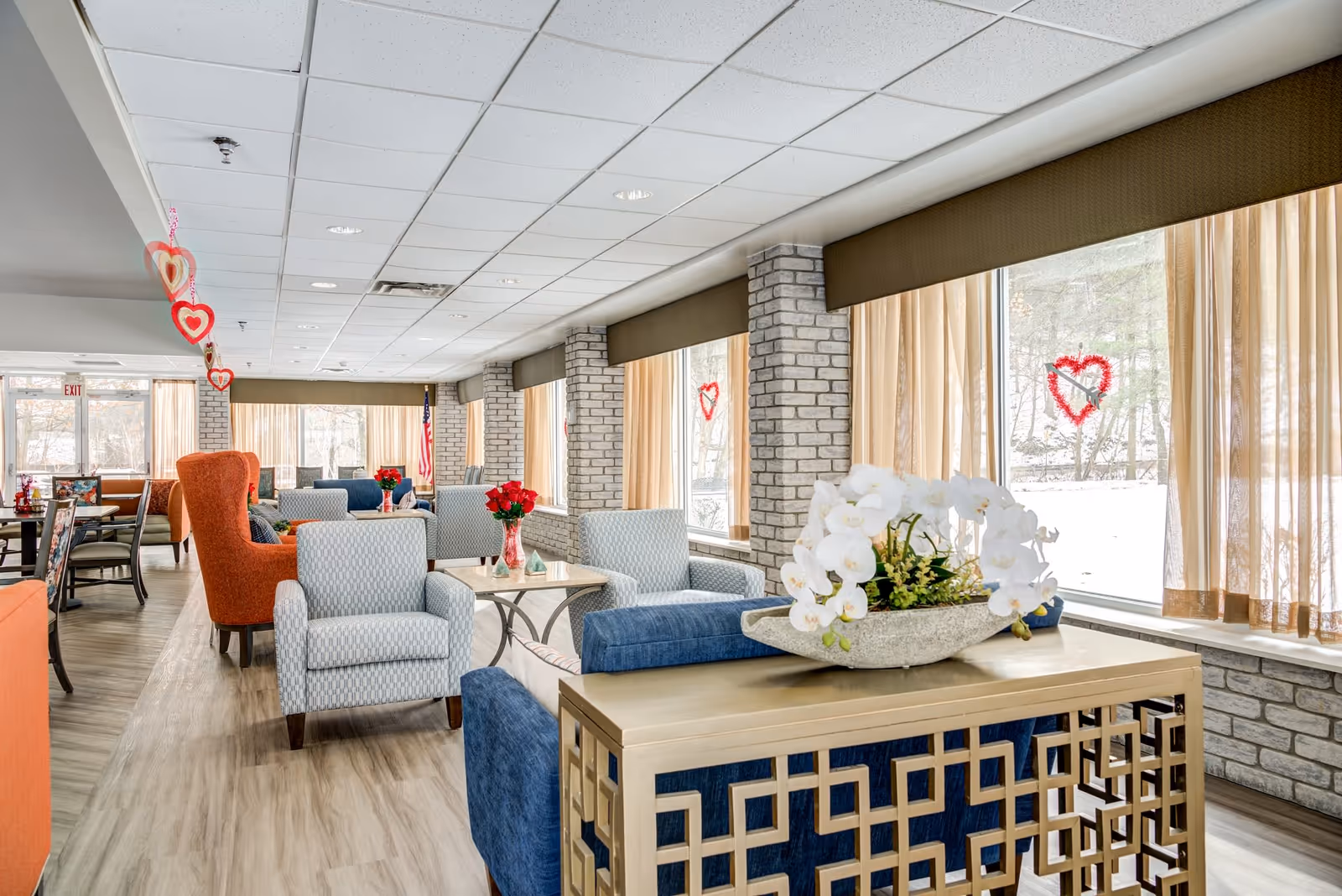 A bright and spacious senior living facility lounge area with large windows covered by sheer curtains, allowing natural light to fill the room. The space features comfortable seating including blue and patterned armchairs, an orange wingback chair, and small tables with red flower vases. Heart-shaped decorations hang from the ceiling and are stuck on the windows. The floor is light wood, and the walls have a light brick finish. An American flag is visible near the back of the room.