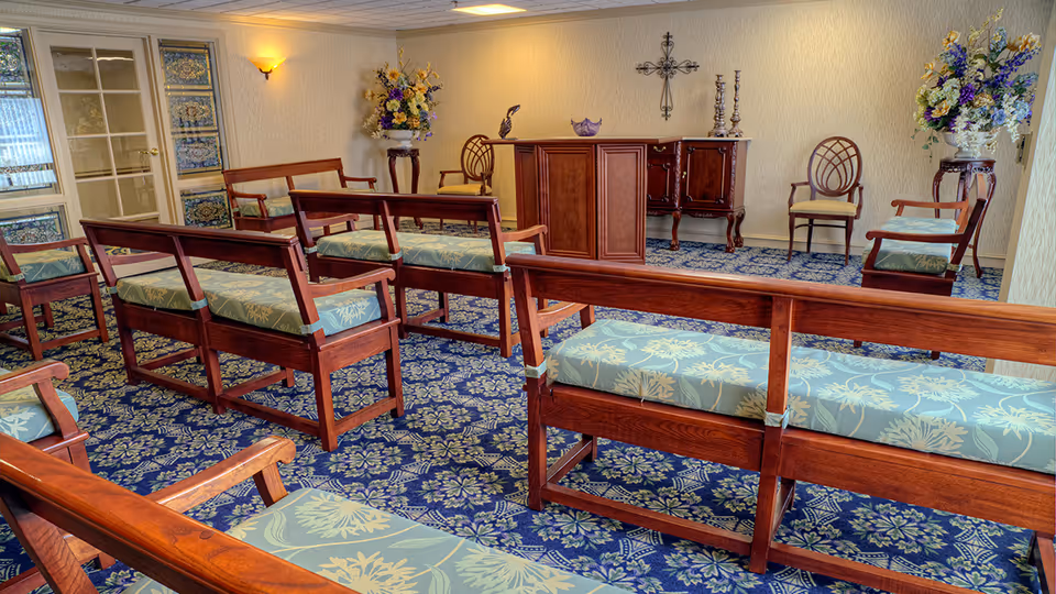 Interior room with wooden benches featuring green floral cushions arranged in rows on a blue patterned carpet. The room has cream-colored walls with a wall-mounted light fixture, floral arrangements on stands, a wooden podium, a sideboard with decorative items, and a cross on the wall.