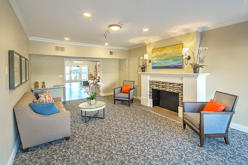 A cozy senior living facility common area with a beige sofa adorned with colorful pillows, two armchairs with orange cushions, a round glass coffee table with a flower arrangement, and a decorative fireplace with a colorful painting above it. The room has patterned carpet flooring and soft lighting from ceiling fixtures and wall sconces.