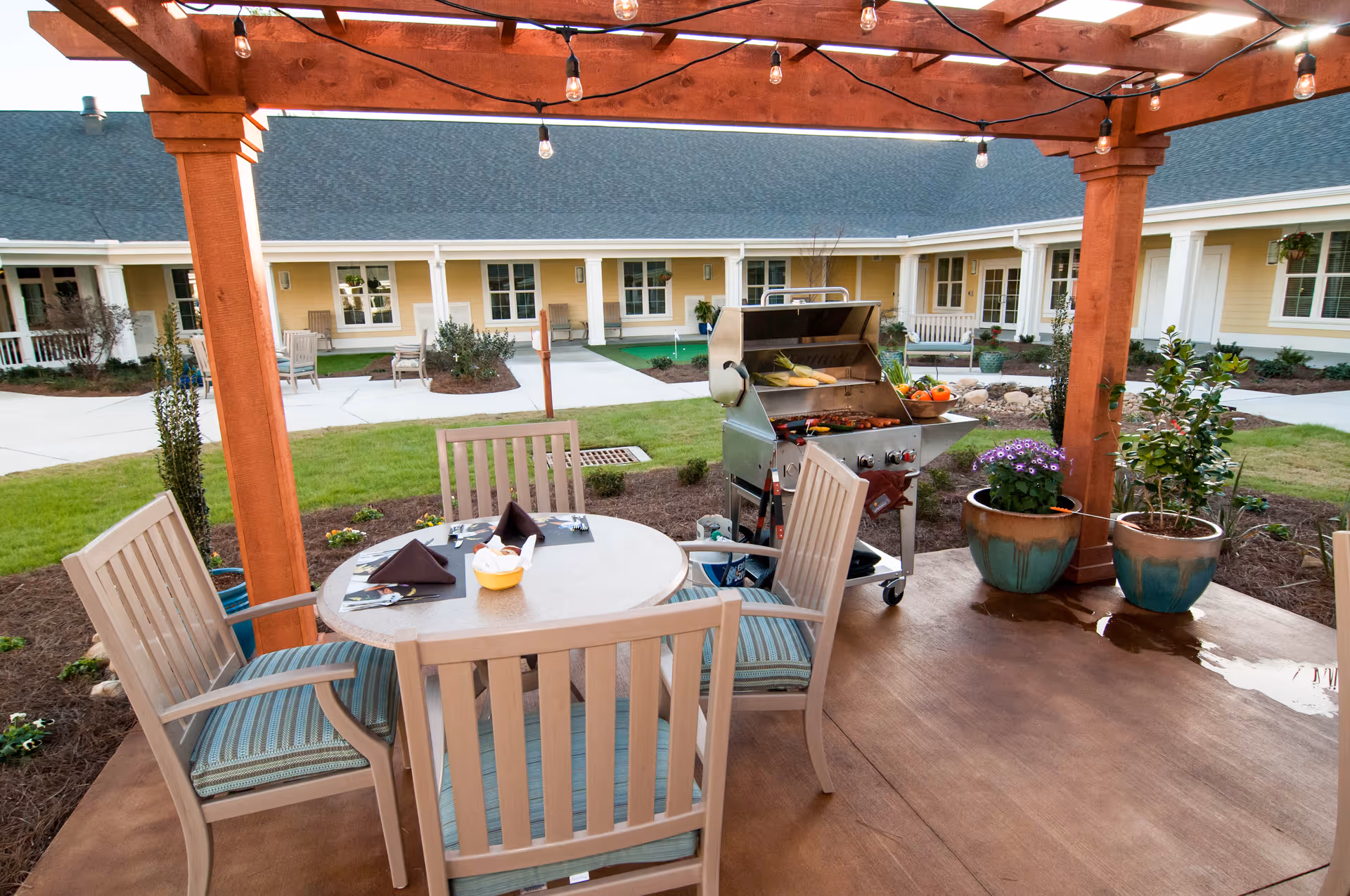 Outdoor patio area with a round table and four cushioned chairs under a wooden pergola adorned with string lights. A grill with food on it is nearby, and potted plants are placed around the patio. In the background, there is a courtyard with grass, walkways, and a building with multiple windows and doors.