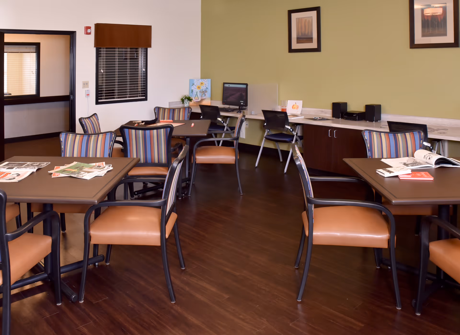 A well-lit room with several tables and chairs arranged for group seating. The chairs have striped backs and tan seats. On the tables are newspapers, magazines, and books. Along one wall, there is a long counter with office chairs and a computer workstation. The walls are painted light green and decorated with framed artwork. The floor is dark wood.