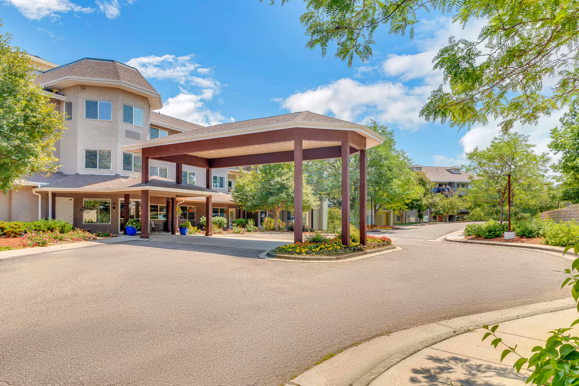 Exterior view of Quincy Place | Sky Active Living facility showing a covered entrance with a driveway, surrounded by trees and landscaping under a partly cloudy sky.