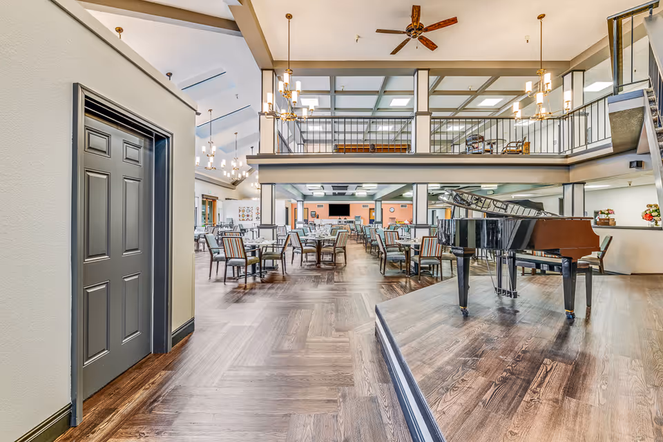 Spacious senior living facility dining area with multiple tables and chairs arranged neatly. A grand piano is placed on a raised platform to the right. The room features high ceilings with chandeliers, a mezzanine level with railings, and large windows allowing natural light.
