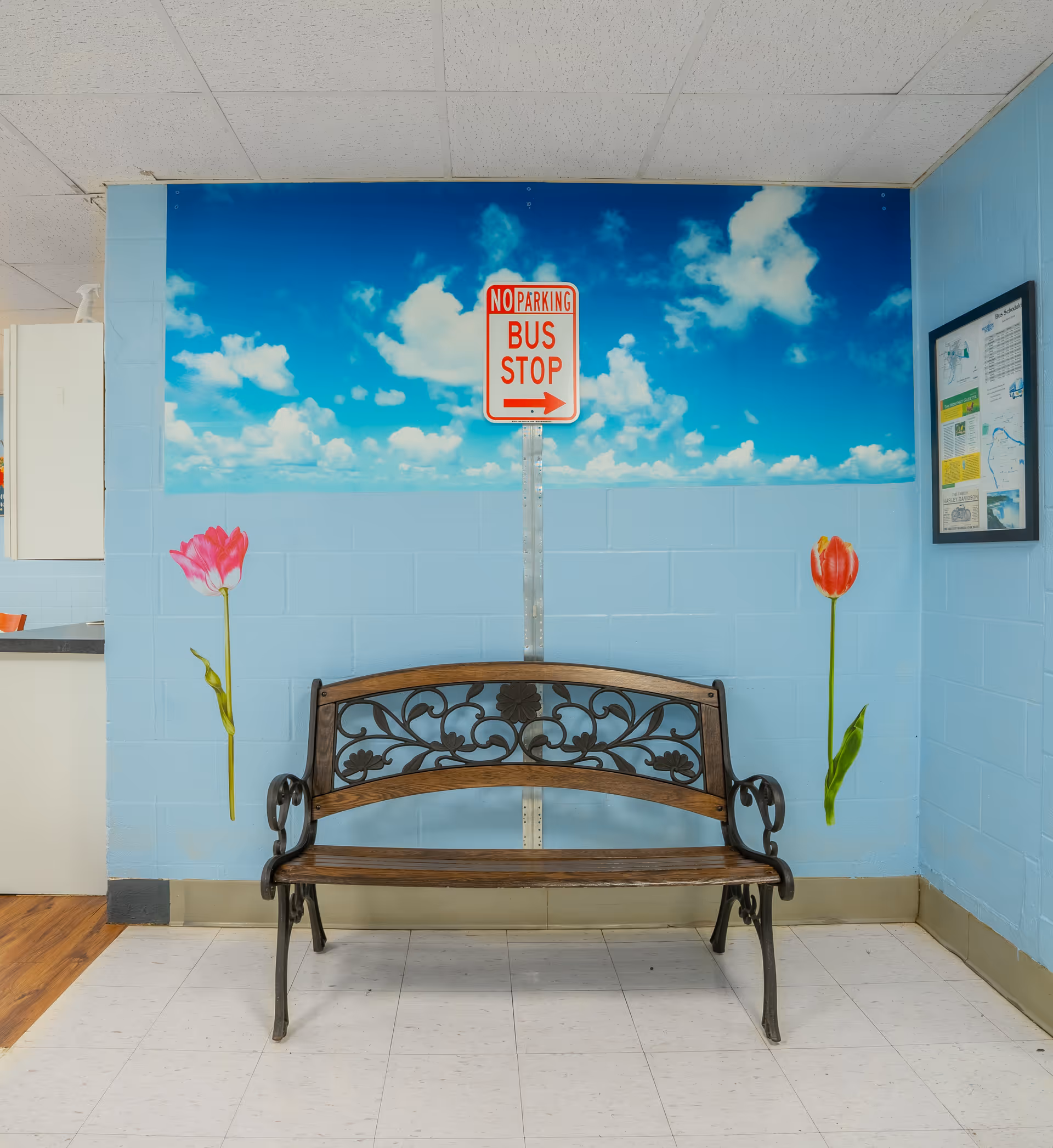 A decorative wooden bench sits against a blue wall painted with a sky mural, a 'No Parking Bus Stop' sign, and two tulip decals.