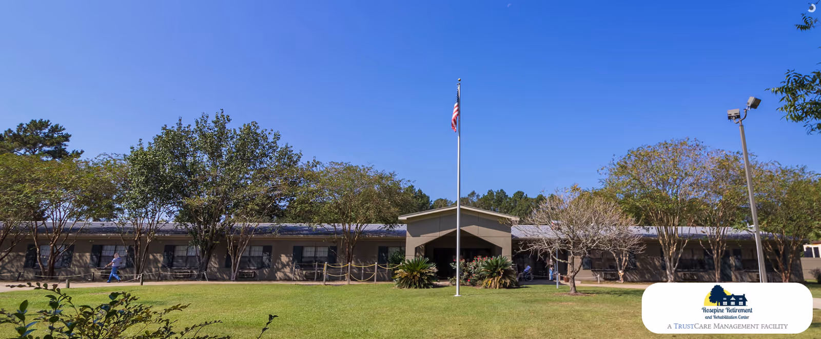 Exterior view of Rosepine Retirement & Rehabilitation Center building with a green lawn, several trees, and an American flag on a flagpole in front under a clear blue sky.