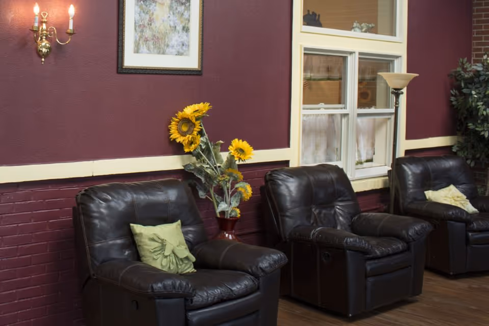 A cozy sitting area with three black leather recliner chairs each with a decorative pillow, a vase with sunflowers on a small table, a floor lamp, a window with white curtains, a framed floral painting on a maroon wall, and a brass wall sconce with two lit bulbs.