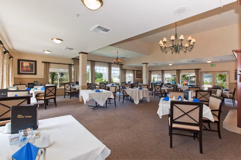 A spacious dining room in Arbor Village Senior Living with multiple tables covered in white tablecloths, each set with blue napkins and dining utensils. The room features large windows with curtains allowing natural light to fill the space, chandeliers hanging from the ceiling, and framed artwork on the walls.