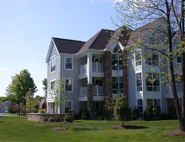 Exterior view of a multi-story residential building with white siding and stone accents, surrounded by green grass, small trees, and a clear blue sky.