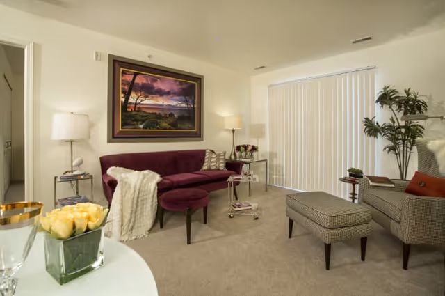 A cozy living room with a purple sofa, a matching ottoman, a patterned armchair with a matching footrest, a side table with a lamp, a small rolling cart, and a large framed landscape painting on the wall. There are vertical blinds covering a window or sliding door, and a potted plant in the corner.