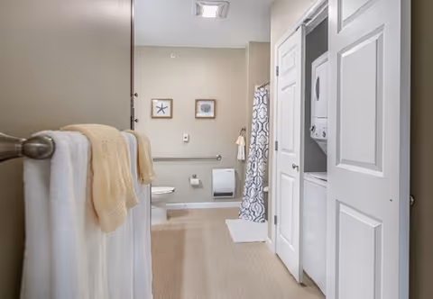Accessible bathroom with towels on a rack, a toilet with grab bar, a shower with a patterned curtain, and a stacked washer-dryer behind bi-fold doors.