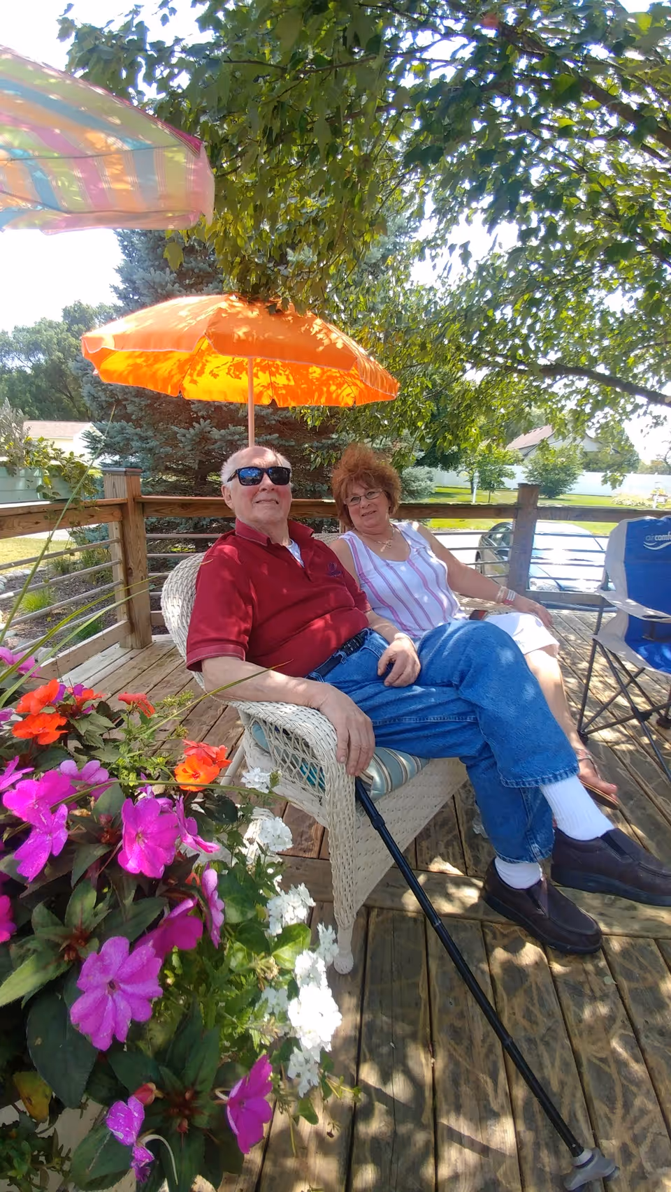 An elderly man and woman sitting together on a wicker chair on a wooden deck under an orange umbrella. The man is wearing sunglasses, a red shirt, blue jeans, and black shoes, holding a cane. The woman is wearing a white and pink striped sleeveless top and white pants. There are colorful flowers in the foreground and trees in the background.