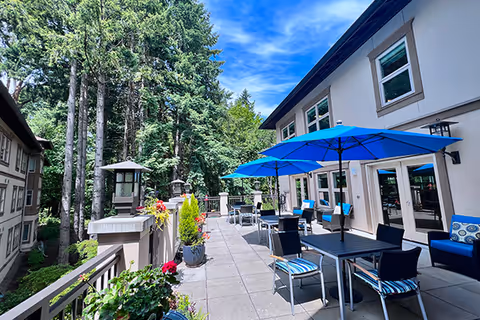 Outdoor patio area at Kellogg Assisted Living At Mary's Woods with tables and chairs under blue umbrellas, surrounded by potted plants and flowers, adjacent to a building with large windows, and tall trees in the background under a partly cloudy blue sky.