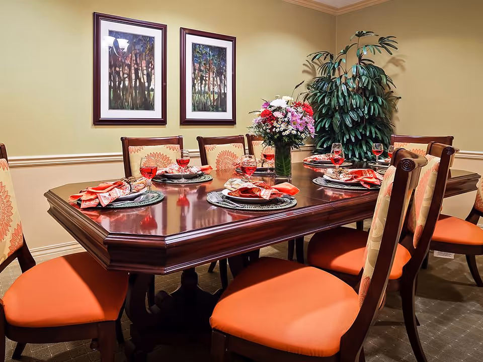 A formal dining room with a dark wooden table set for six people. Each place setting has a woven placemat, a plate with a folded red napkin, and a glass of red beverage. The chairs have wooden frames with orange cushioned seats and patterned backs. On the table is a vase with a colorful bouquet of flowers. The walls are light yellow with two framed paintings and a large green potted plant in the corner.
