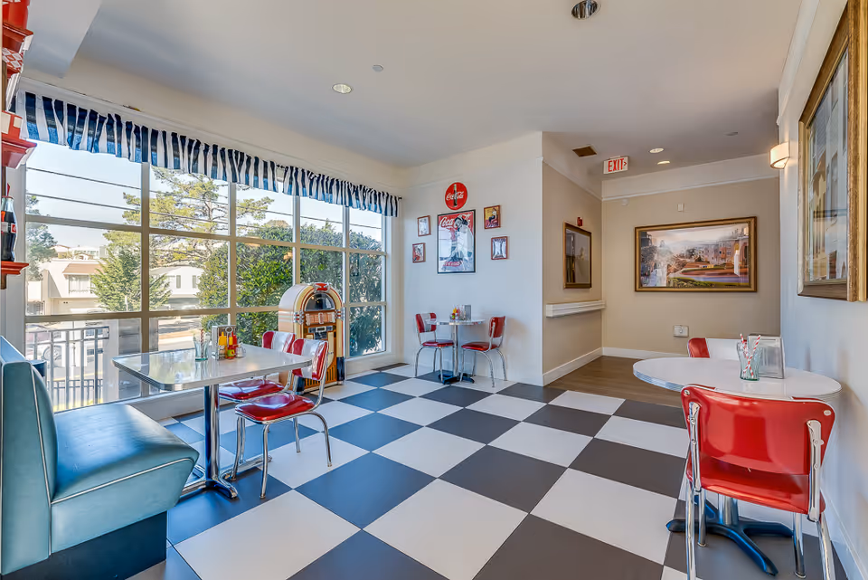 A bright dining area with large windows letting in natural light, featuring a black and white checkered floor, red chairs, a blue bench, and small round and square tables. The walls are decorated with vintage Coca-Cola posters and a jukebox is placed near the window. There are framed pictures on the walls and an exit sign above a hallway.