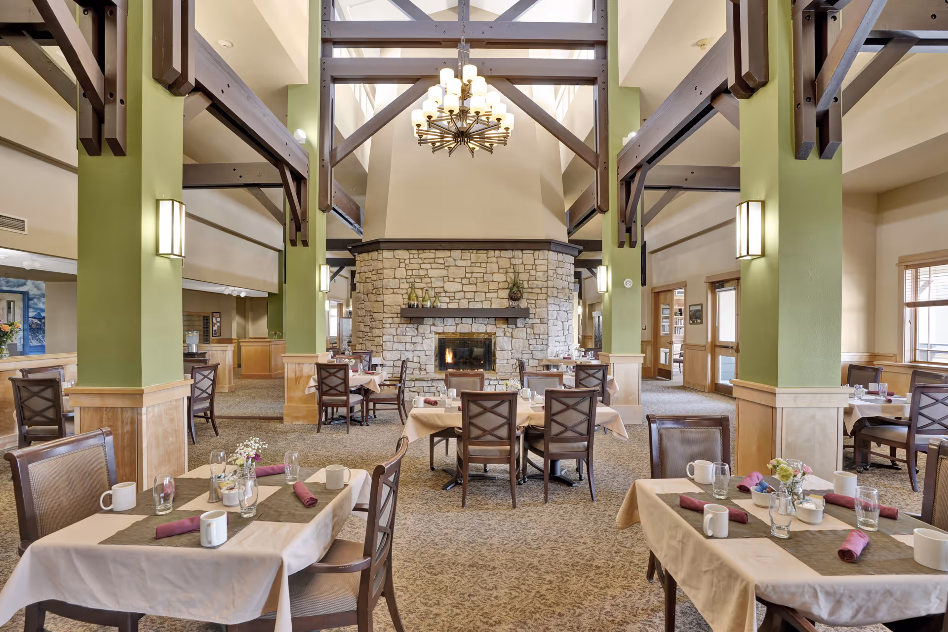 A spacious dining room with multiple tables set with cups, glasses, napkins, and small flower arrangements. The room features green pillars, wooden beams, a large stone fireplace in the center, and a chandelier hanging from the high ceiling. Large windows allow natural light to fill the space.