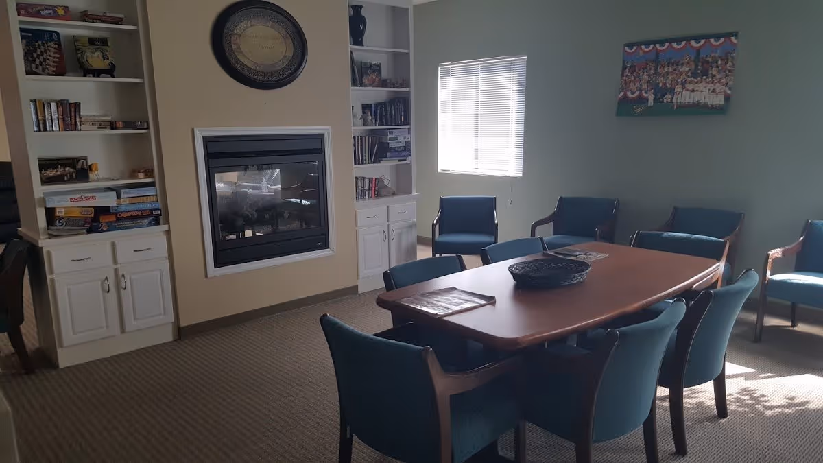A cozy room with a rectangular wooden table surrounded by eight blue upholstered chairs. The room has a beige carpet and light-colored walls. There is a fireplace built into the wall with white cabinetry and shelves on either side, containing books and board games. A window with blinds lets in natural light, and a framed picture hangs on the wall.