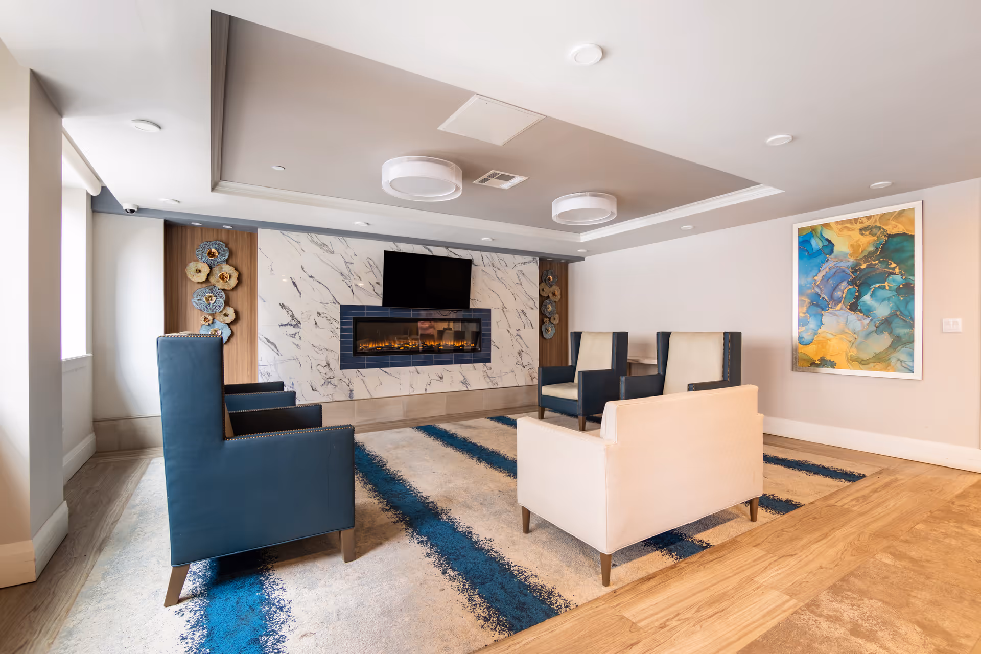 A modern living room area with a marble fireplace and a mounted flat-screen TV above it. The room features four armchairs arranged around a blue and beige striped rug. There is a colorful abstract painting on the wall to the right and decorative wall art on the left side of the fireplace. The ceiling has recessed lighting and two circular light fixtures.