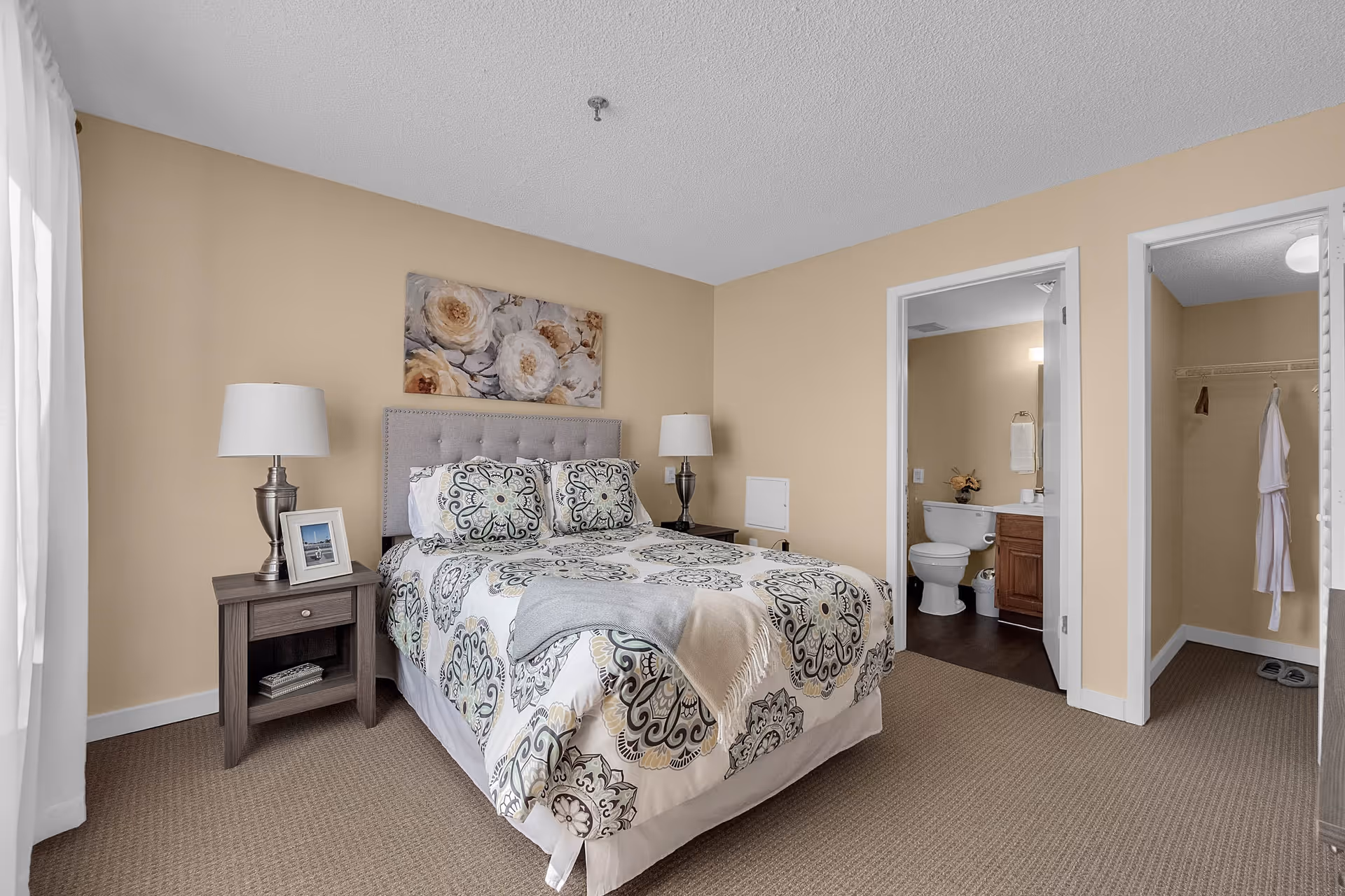 A bedroom in a senior living facility with a bed featuring patterned bedding and two pillows. There are two bedside tables each with a lamp, and a framed photo on the left table. The walls are beige, and there is a floral painting above the bed. To the right, there are two open doorways leading to a bathroom with a toilet and vanity, and a walk-in closet with a hanging robe and slippers on the floor.