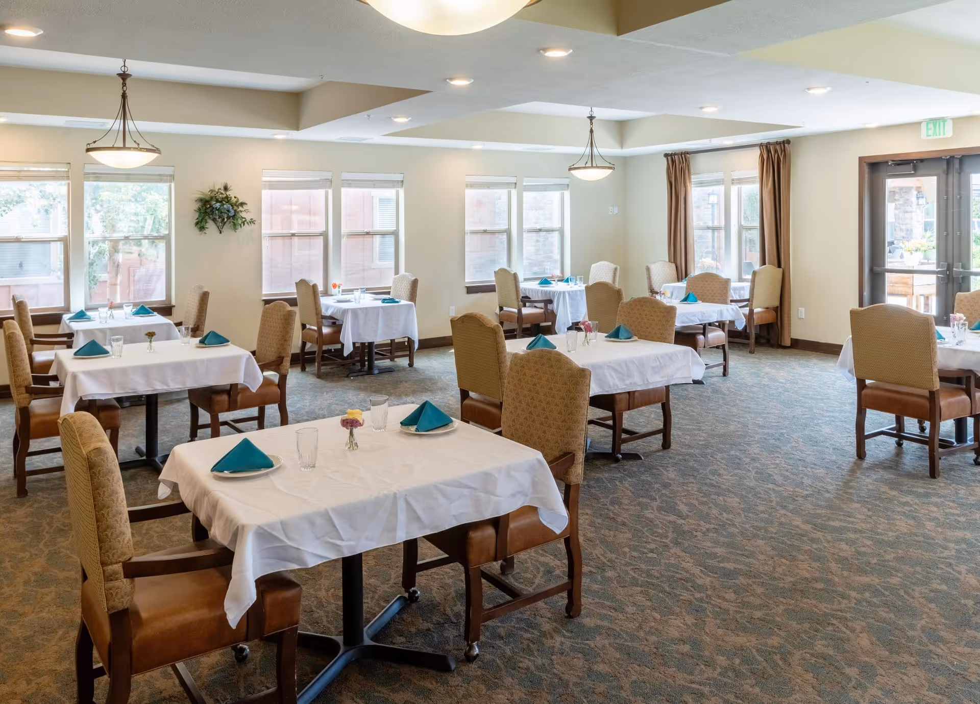 Dining room with multiple tables set with white tablecloths and teal napkins, upholstered chairs, and large windows.
