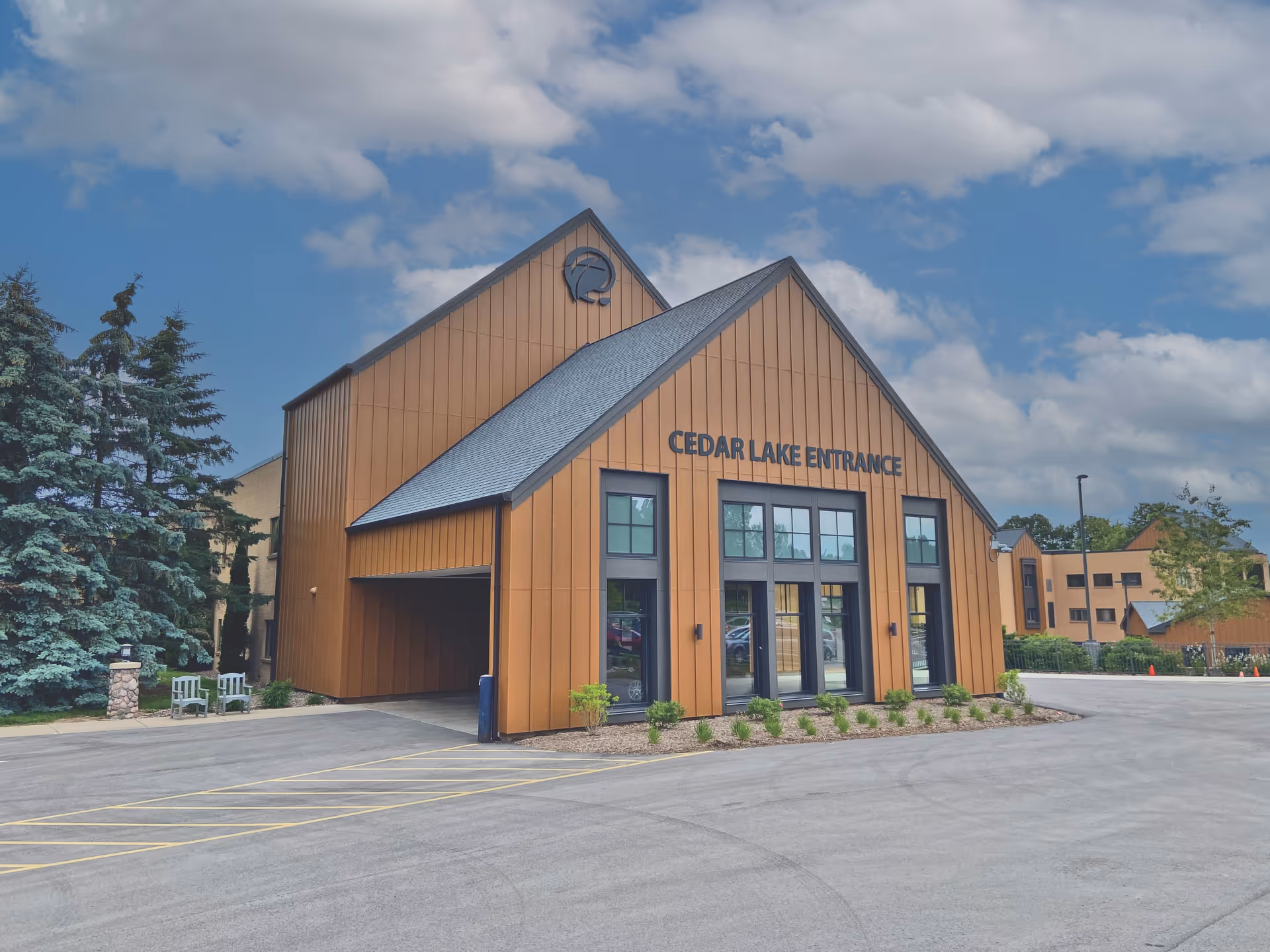 Front exterior of the Cedar Lake Entrance building with wood-paneled siding, large windows, and a paved driveway under a cloudy sky.