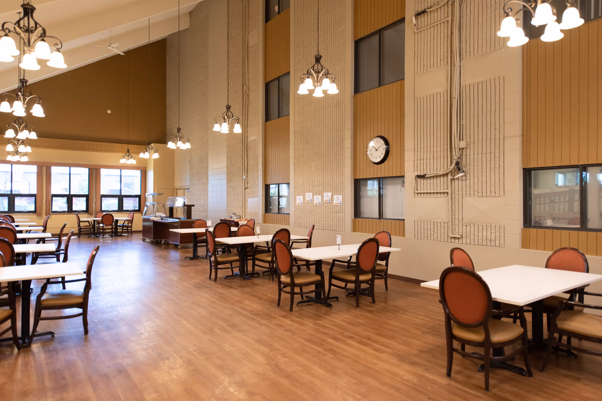 A spacious dining room with multiple tables and chairs arranged neatly on a wooden floor. The room has high ceilings with several hanging light fixtures, large windows letting in natural light, and a clock on the wall. The walls are a combination of beige and brown tones.