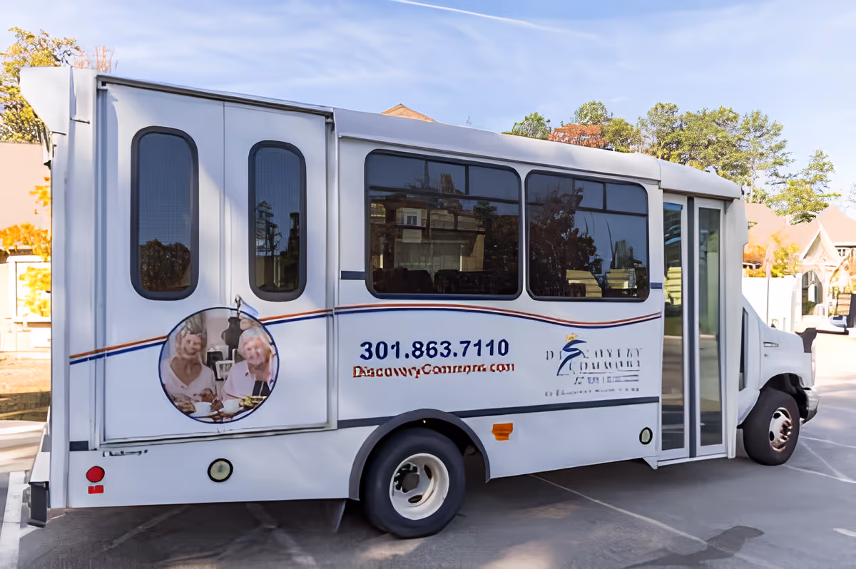 A white shuttle bus parked outdoors with windows and a sliding door. The side of the bus displays the phone number 301.863.7110 and the website DiscoveryCommons.com, along with the Discovery Commons logo and an image of two elderly women smiling and enjoying a meal.