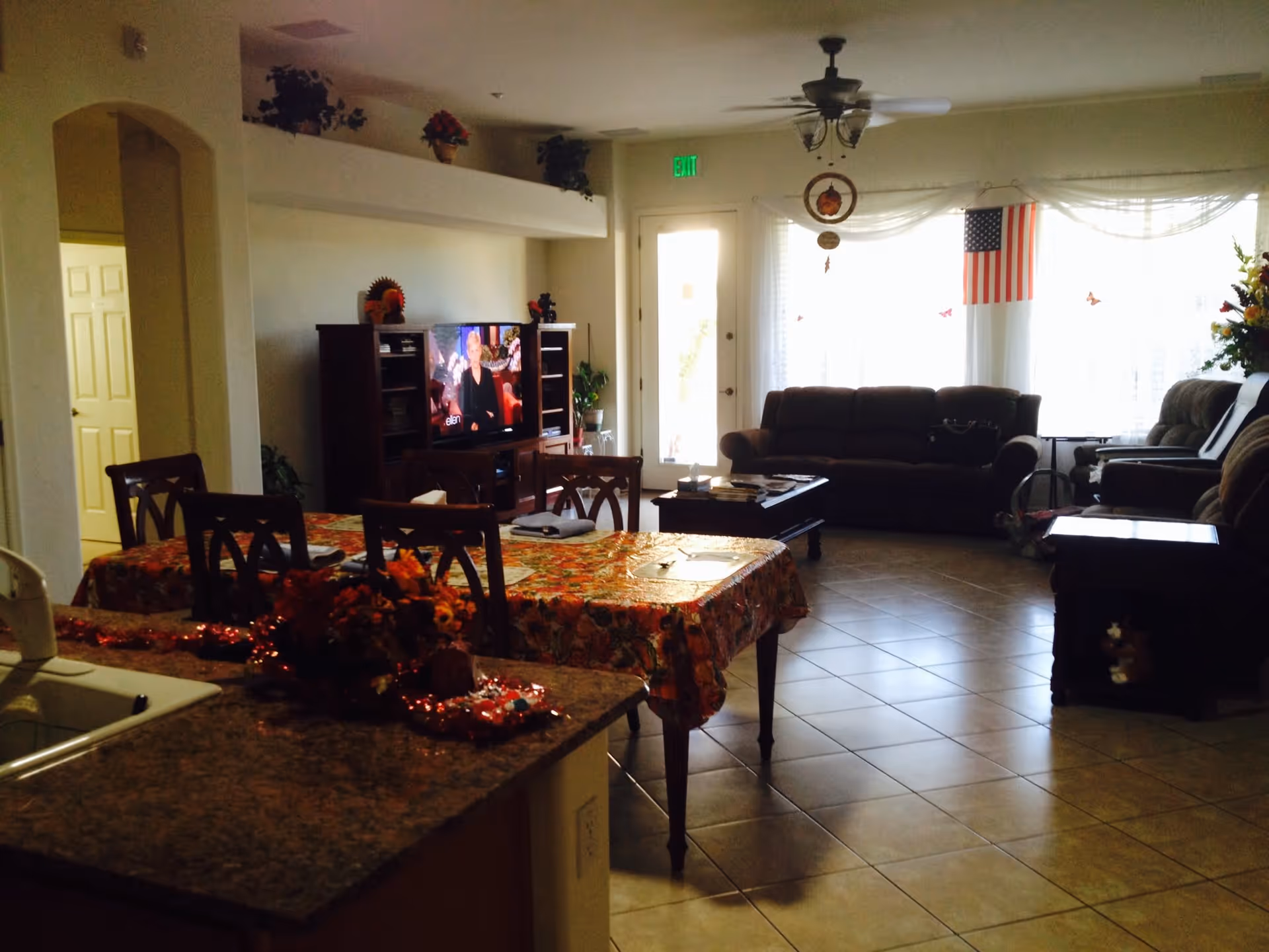 A cozy living room and dining area in a senior living facility with a table covered in a floral tablecloth, several chairs, a TV on a wooden stand, a brown sofa, and an American flag hanging near a window with sheer curtains. The room has tiled floors and some decorative plants and flowers.