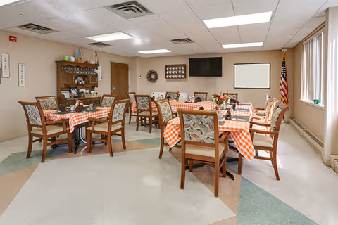 A dining room with several wooden tables covered with orange and white checkered tablecloths. Each table is surrounded by wooden chairs with floral patterned cushions. The room has beige walls, a tiled floor with a geometric pattern, and fluorescent ceiling lights. There is a wooden hutch with decorative items, a wall-mounted TV, an American flag in the corner, and a window with vertical blinds letting in natural light.