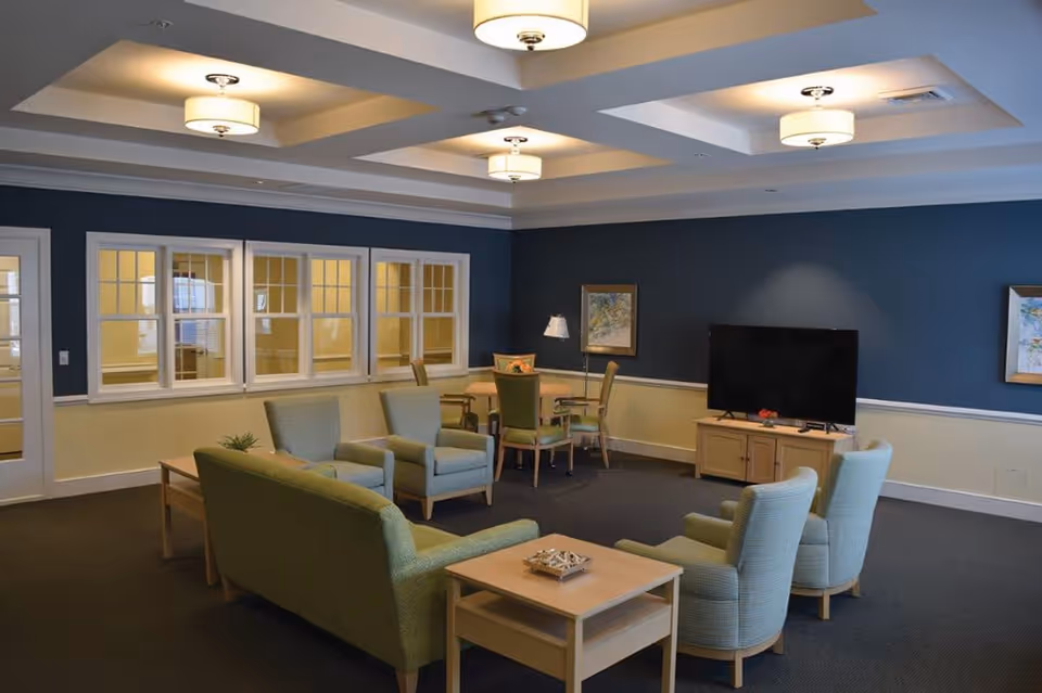 A spacious living room with a green sofa, four armchairs, and a wooden coffee table in the center. There is a TV on a wooden stand against a blue accent wall, and a small dining table with four chairs in the corner. The room has multiple ceiling lights and windows with white frames along one wall.