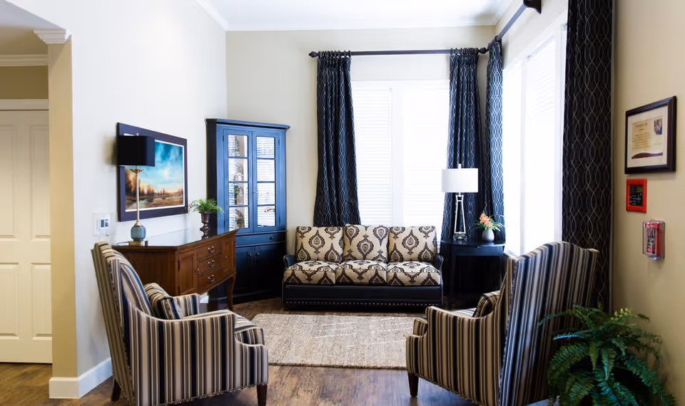 A cozy living room area with a patterned sofa, two striped armchairs, a wooden cabinet with a lamp and plant, a black display cabinet, and large windows with dark curtains letting in natural light. The room has wooden flooring and light-colored walls.