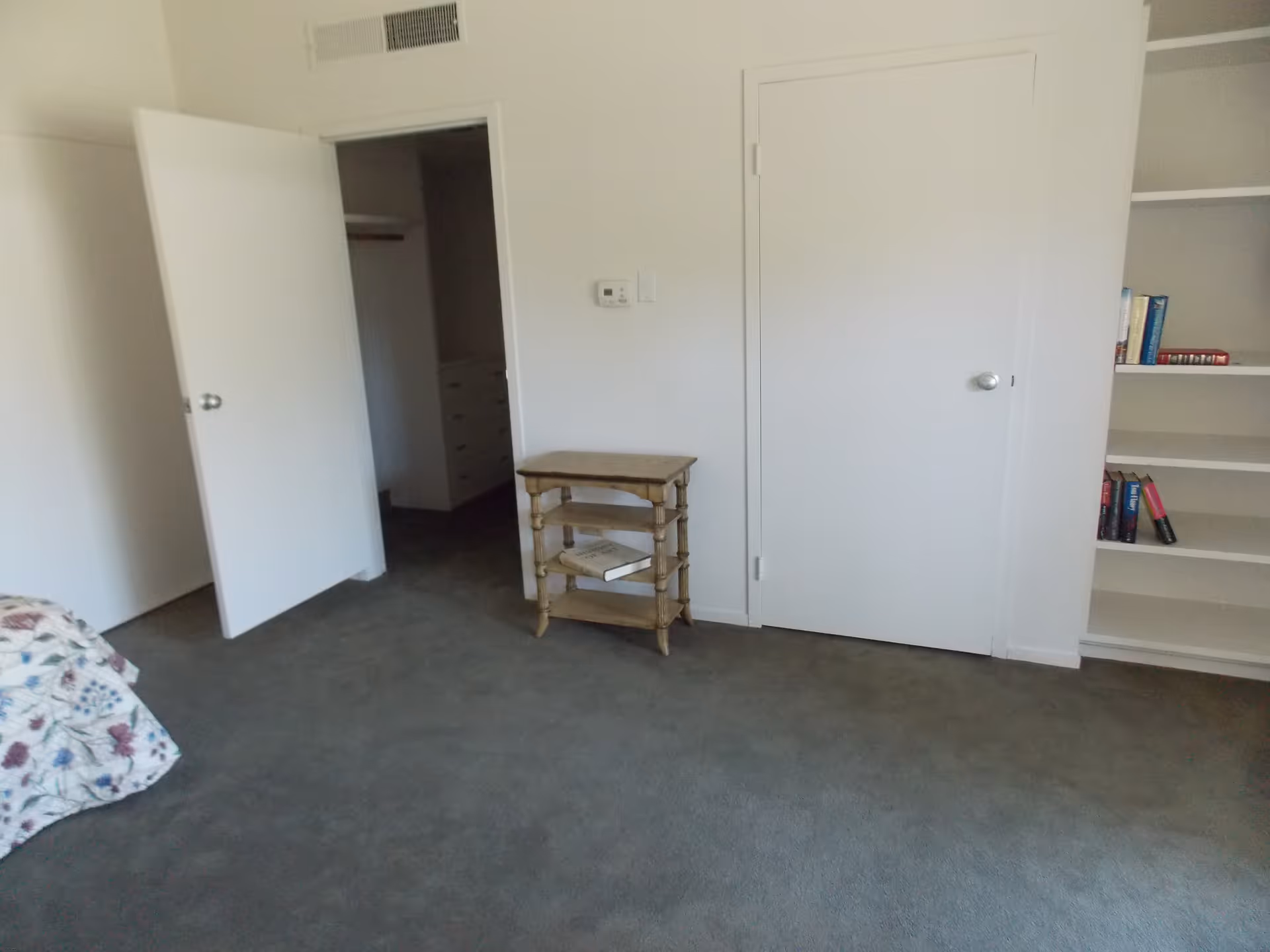 A room with gray carpet, white walls, and two closed white doors. One door is open, revealing a closet with drawers inside. There is a small wooden side table with two books on the lower shelves, and a built-in white bookshelf with some books on the right side. Part of a bed with a floral bedspread is visible on the left edge of the image.