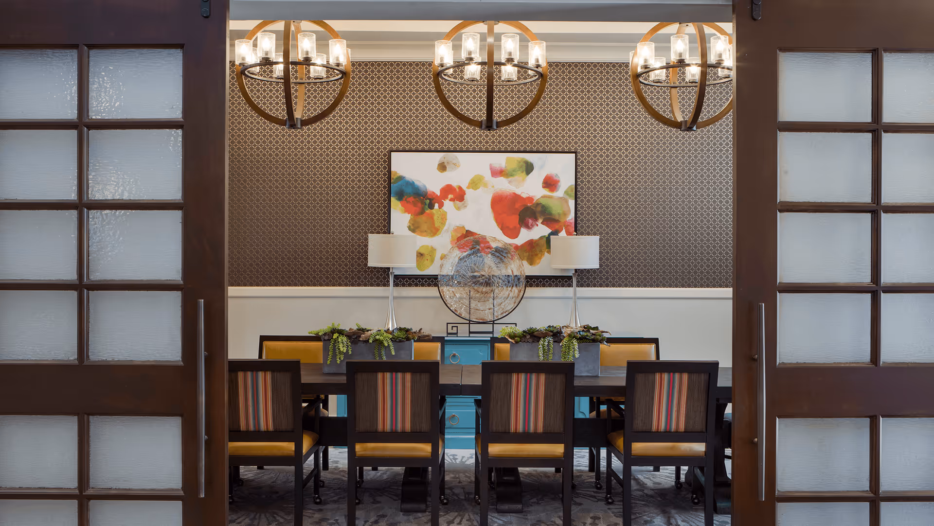 A dining room with a long dark wooden table surrounded by chairs with striped backs and yellow seats. The room features three modern circular chandeliers hanging from the ceiling, a patterned wallpaper on the upper half of the wall, and a colorful abstract painting above a blue sideboard with two lamps and decorative plants.