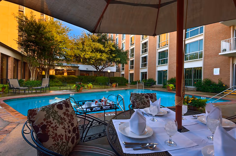 Outdoor dining table set under an umbrella beside a swimming pool in a courtyard surrounded by a multi-story brick building.