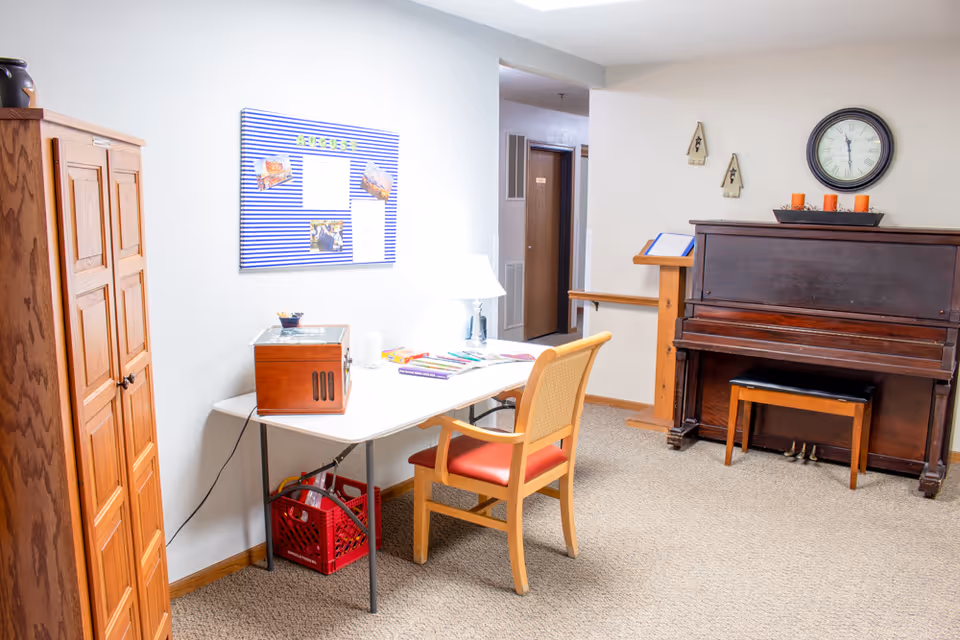 Small activity room with a table and chair, upright piano, bulletin board and wooden cabinet.