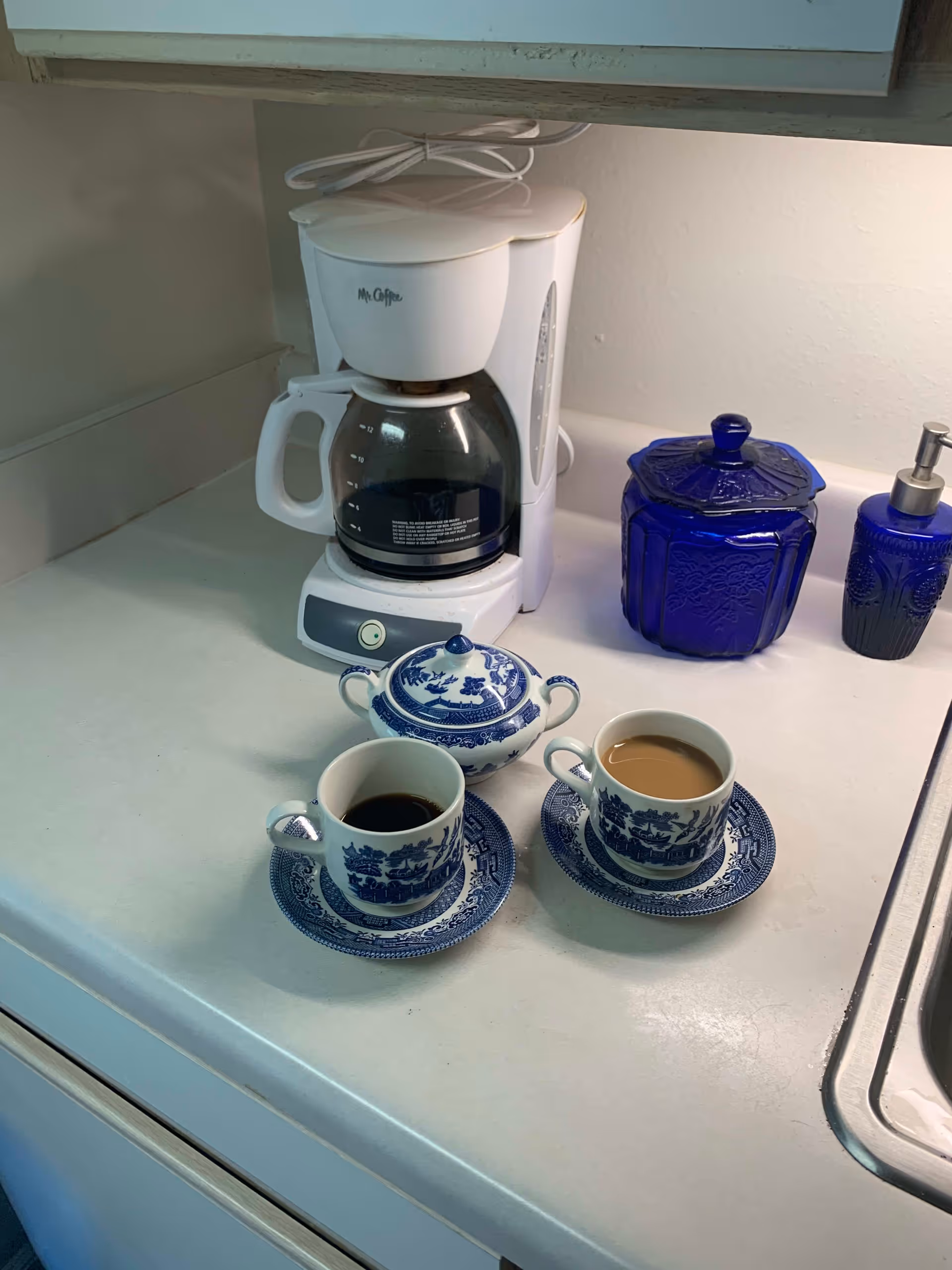 A white Mr. Coffee coffee maker on a kitchen counter with two blue and white patterned cups and saucers filled with coffee, a matching sugar bowl with a lid, and two blue containers, one with a pump dispenser, next to a kitchen sink.