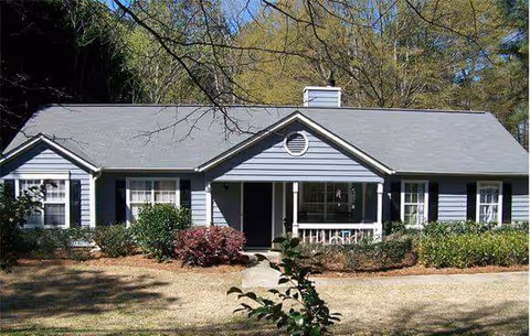 Single-story light-blue house with a central front porch, shutters, shrubs, and a lawn under trees.