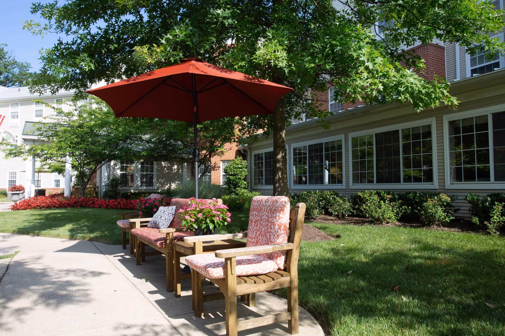 Outdoor seating area at Morningside House of Satyr Hill with wooden chairs and benches featuring patterned cushions, a red umbrella providing shade, surrounded by green grass, trees, and flowering plants near a building with large windows.