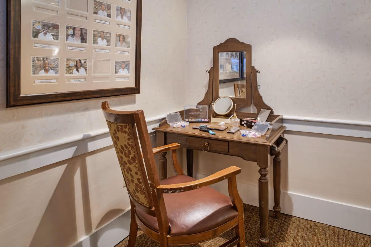 A wooden vanity table with a mirror, makeup items and a round magnifying mirror, and a patterned wooden chair set against a wallpapered wall beneath a framed staff photo collage.