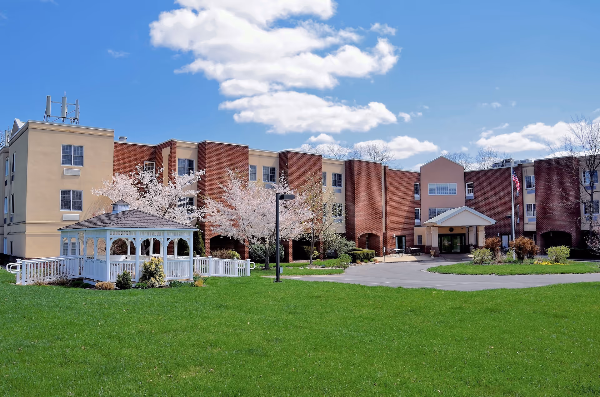 Exterior view of a senior living facility building with a mix of beige and red brick walls under a partly cloudy blue sky. In front of the building is a well-maintained green lawn with a white gazebo and some blossoming trees. A paved driveway leads to the building entrance, which has a covered porch and an American flag on a flagpole nearby.