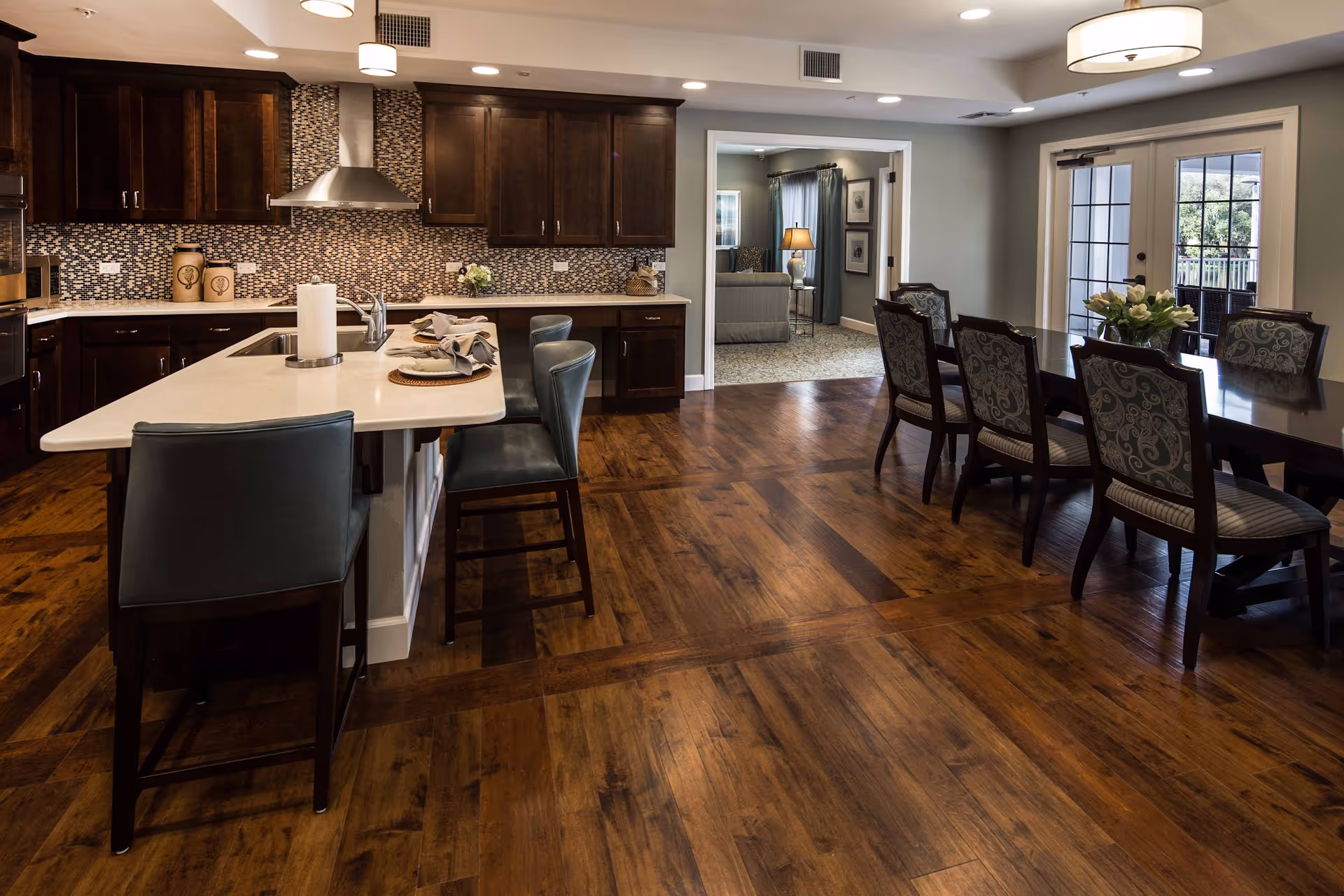A spacious kitchen and dining area with dark wooden cabinets, a white countertop island with bar stools, a dining table with six upholstered chairs, wooden flooring, and a doorway leading to a living room and an outdoor patio.