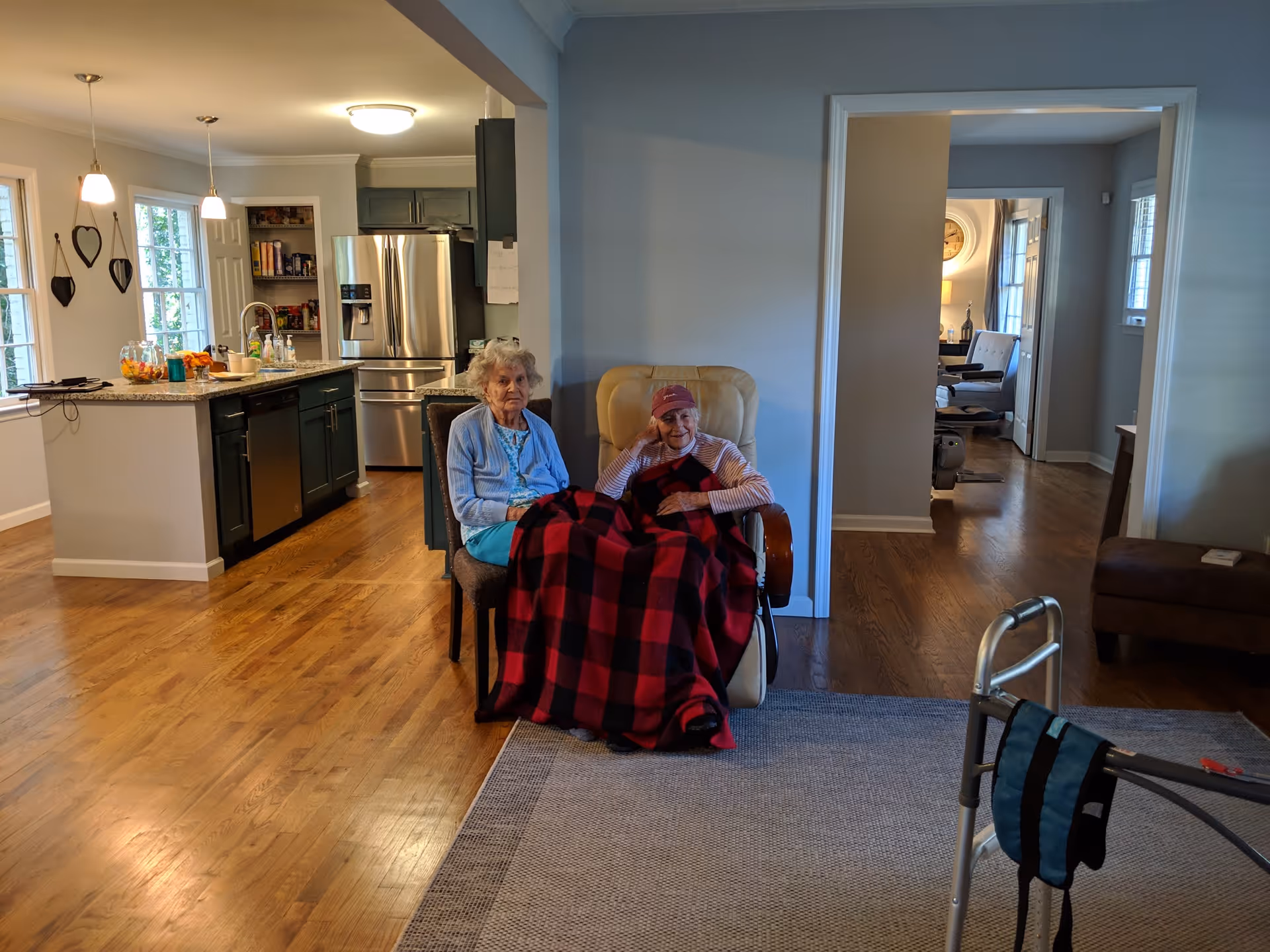 Two elderly women sitting together in chairs in a cozy living area with a red and black checkered blanket covering their laps. The room has wooden floors and is adjacent to a modern kitchen with stainless steel appliances and pendant lighting. A walker is visible in the foreground on a gray rug.
