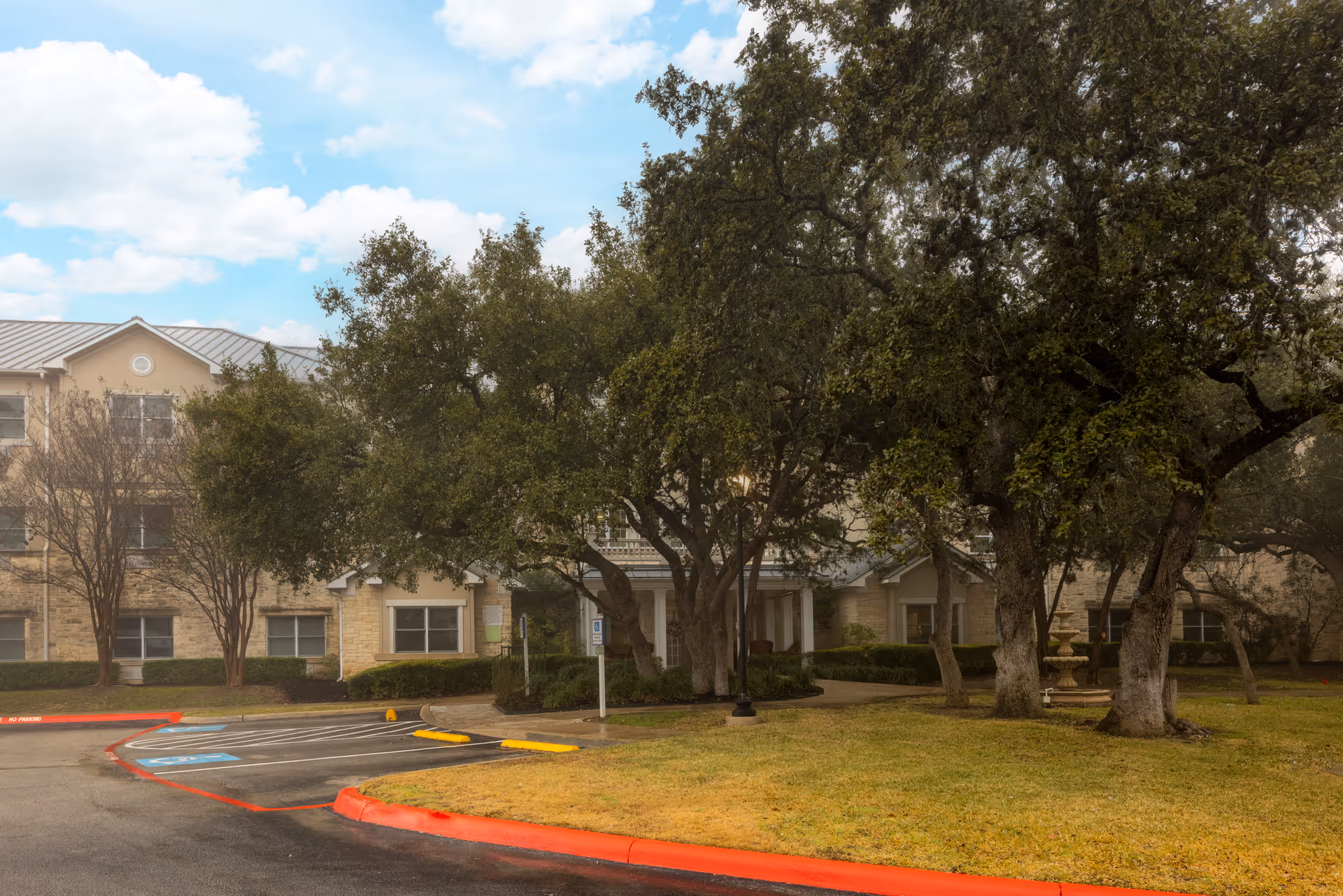 Exterior view of a senior living facility building with a beige stone facade, surrounded by large trees and a grassy lawn. There is a parking area with handicap parking spaces and a red curb in the foreground under a partly cloudy sky.