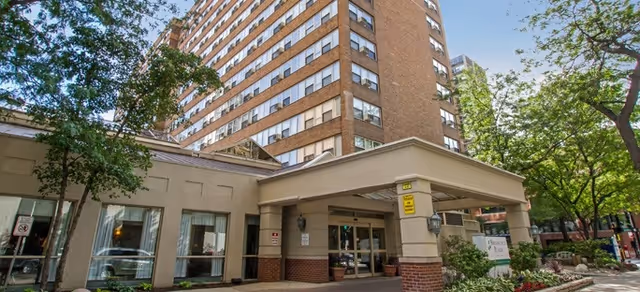 Covered entrance and landscaped front of a multi-story brick senior living building under a blue sky.