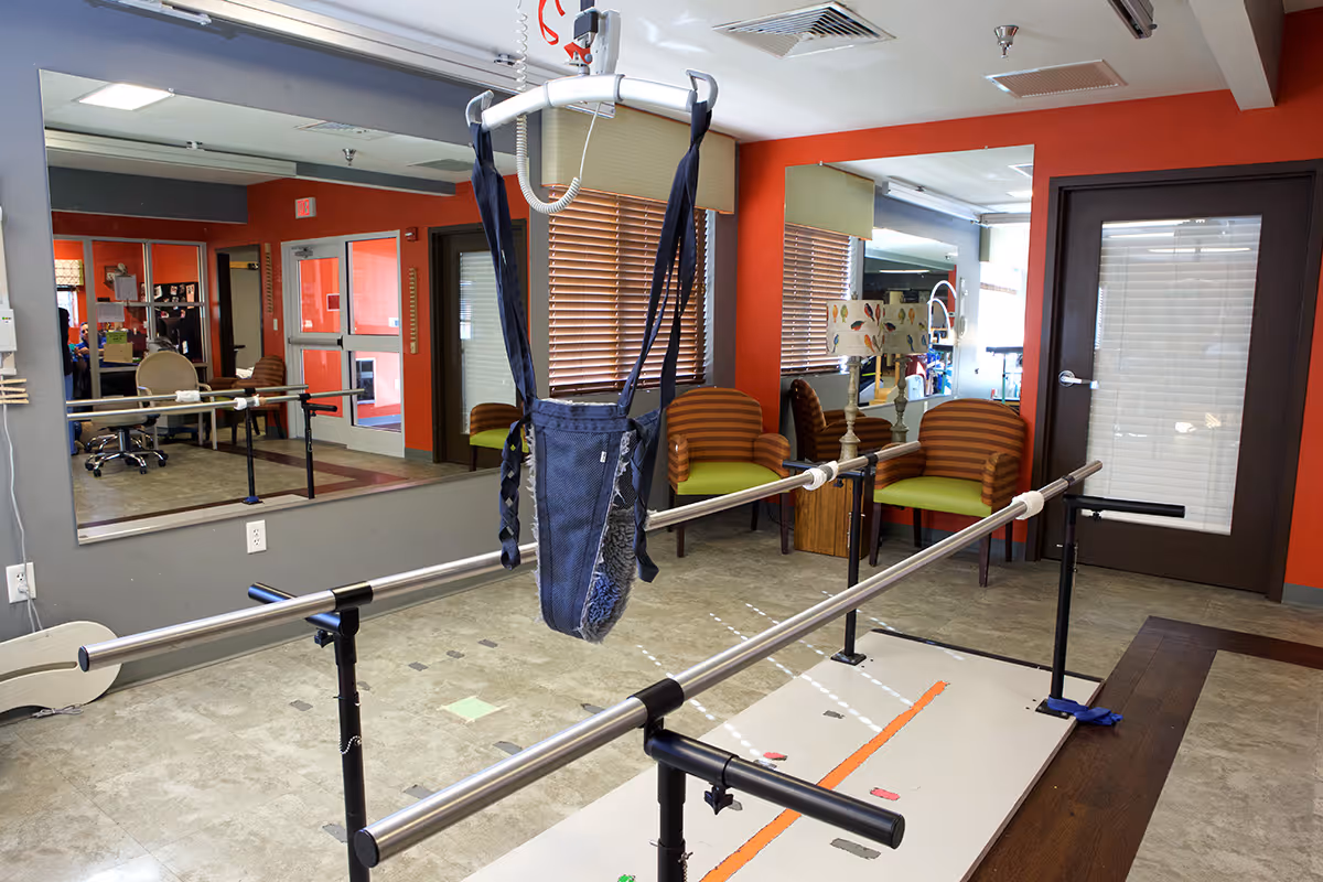 Physical therapy room with parallel bars for walking exercises, a patient lift sling hanging from the ceiling, two striped chairs with green cushions, a floor lamp with a bird-patterned shade, large wall mirrors, and a door with a window and blinds.