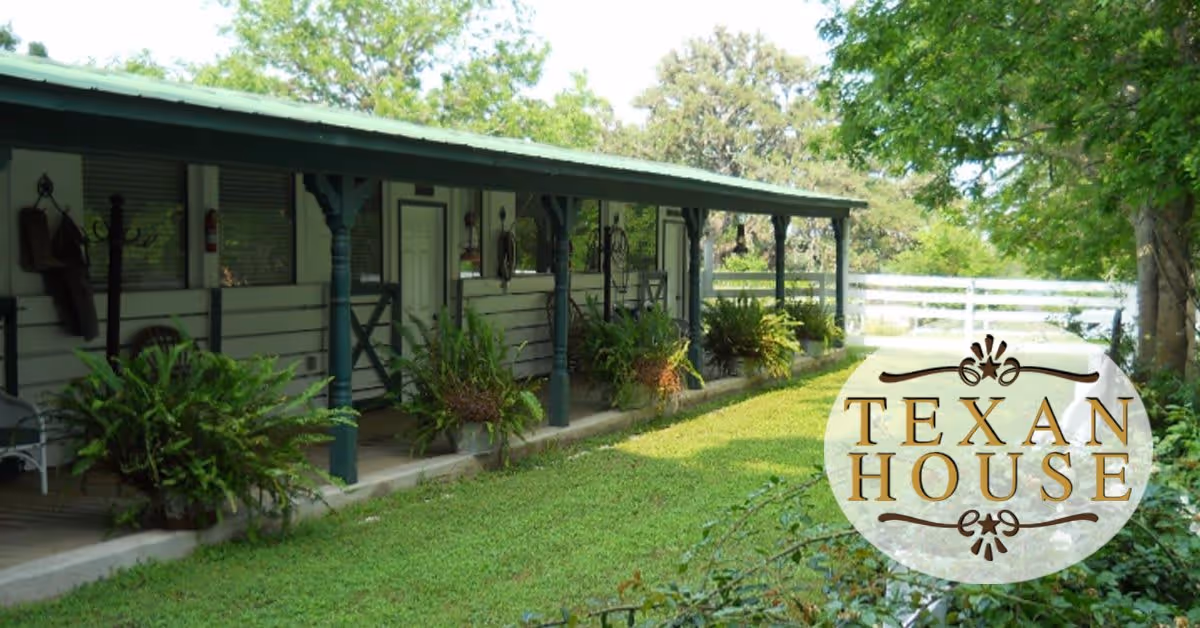 Exterior view of a single-story building with a covered porch supported by dark green pillars. Several doors and windows line the porch, with hanging plants and ferns placed along the edge. The building is surrounded by green grass and trees, and a white fence is visible in the background. The image includes a logo that reads 'Texan House'.
