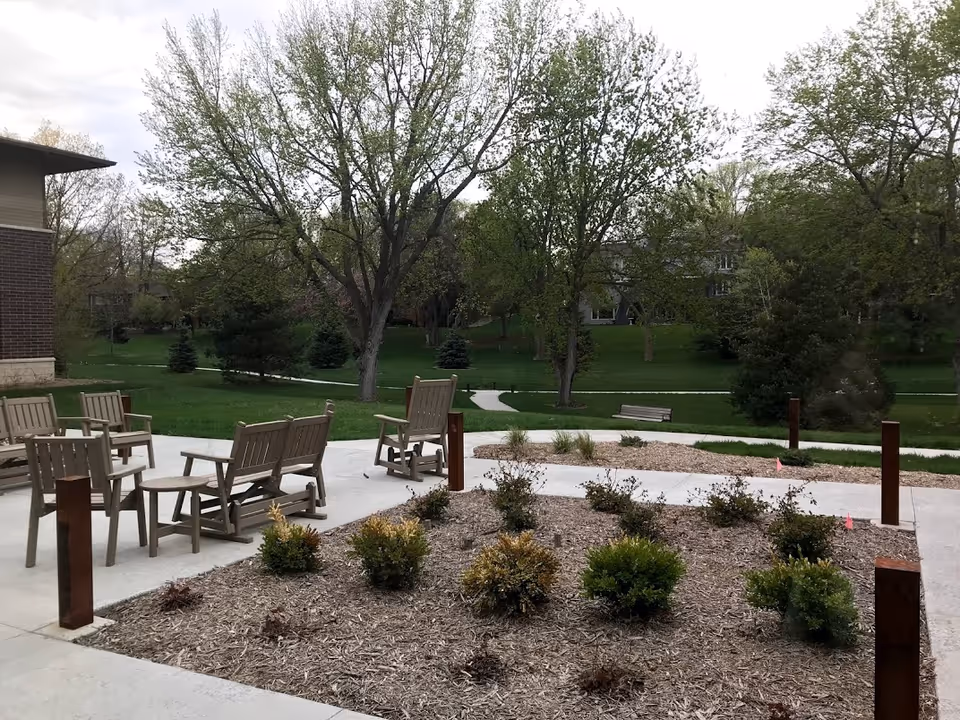 Outdoor patio area with wooden chairs and benches arranged around small tables, surrounded by landscaped garden beds with shrubs and trees in the background, part of a senior living facility.