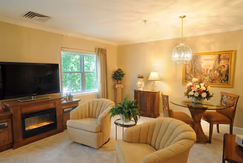 A cozy living room with two beige upholstered armchairs, a wooden entertainment center with a flat-screen TV and electric fireplace, a window with beige curtains, a small round glass dining table with four chairs, a floral centerpiece on the table, a sideboard with two lamps and decorative items, a potted plant on a pedestal, and a framed painting on the wall.